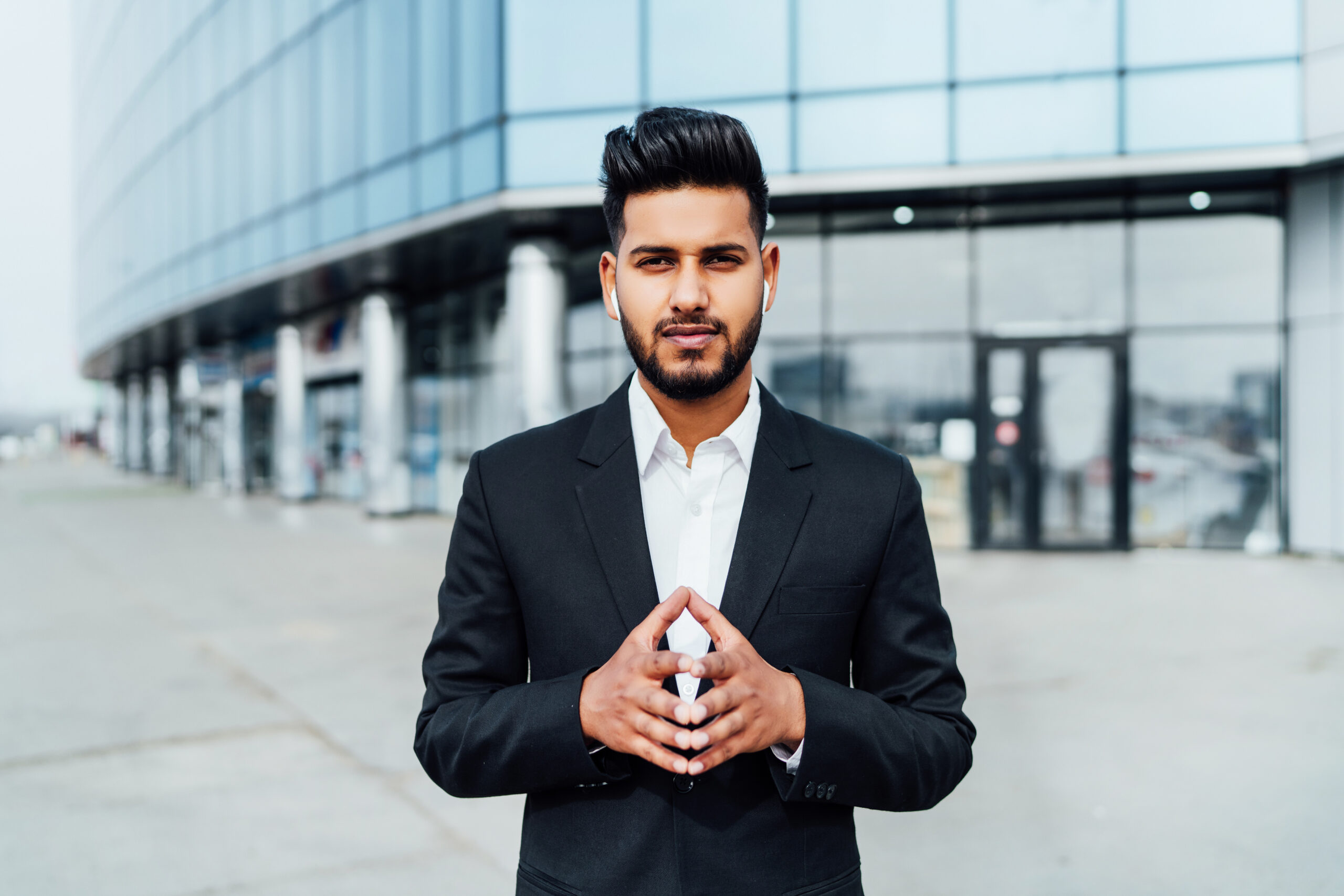 Portrait of a serious smiling modern Indian man, businessman, in front of the holding, behind him a modern building, he is looking at the camera.